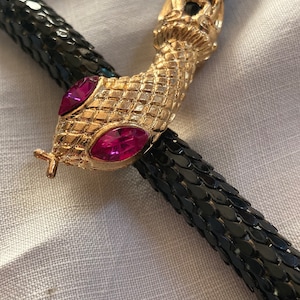 Może przedstawiać: Czarny pasek ze złotą klamrą w kształcie głowy węża. Głowa węża ma oczy z różowych kamieni szlachetnych. Pasek wykonany jest z małych, zachodzących na siebie czarnych łusek.