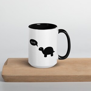 May include: White ceramic coffee mug with a black rim and handle. The mug features a black silhouette of a turtle with a speech bubble that says "cluck".