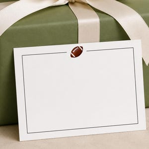 May include: A rectangular white note card featuring a thin black border and a small brown football icon centered at the top, positioned in front of a square gift box wrapped in olive green paper and tied with a wide cream-colored satin ribbon.