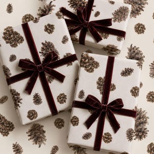 Può includere: Tre scatole regalo rettangolari avvolte in carta color bianco sporco con un motivo ripetuto di pigne marroni. Ogni pacchetto è legato con un nastro di velluto bordeaux scuro disposto a croce e rifinito con un fiocco centrale.
