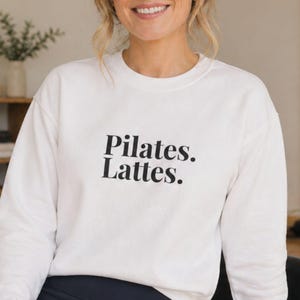 May include: White crew neck sweatshirt with the text "Pilates. Lattes." in black. The sweatshirt is made of a soft material and is suitable for casual wear. The design is simple and modern, perfect for fitness enthusiasts.