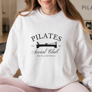 Peut inclure: Sweat-shirt blanc avec un motif noir "Pilates Social Club". Le motif comprend le mot "PILATES" au-dessus d'une illustration d'un appareil de Pilates, avec le texte "Social Club" et "Meet You at the Reformer" en dessous.