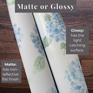 May include: Two rolls of wallpaper featuring a blue hydrangea floral pattern. Text labels indicate Matte or Glossy finishes, explaining that Matte has a non-reflective flat finish and Glossy has a light catching surface for the wallpaper.