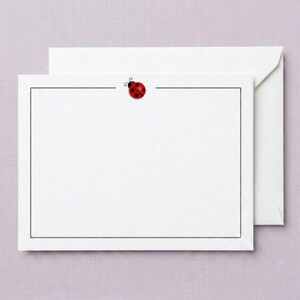 May include: A white rectangular note card featuring a thin black border and a small red ladybug with black spots centered at the top edge. The card is accompanied by a matching white envelope positioned directly behind the note card.