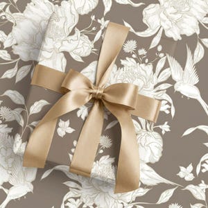 May include: A rectangular gift box wrapped in taupe paper featuring a white line-art pattern of blooming peonies and small birds. The box is tied with a wide, smooth, metallic gold satin ribbon arranged in a large, centered decorative bow.