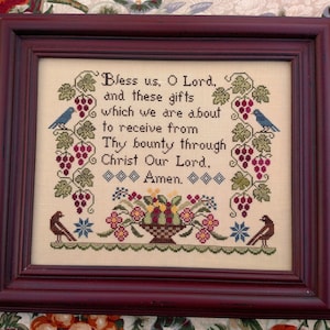 May include: A framed cross-stitch embroidery featuring a traditional Thanksgiving prayer. The prayer reads "Bless us, O Lord, and these gifts which we are about to receive from Thy bounty through Christ Our Lord, Amen."