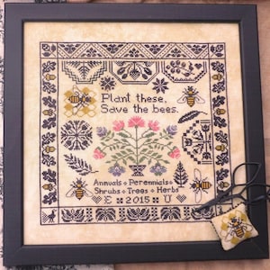 Puede incluir: Arte de punto de cruz enmarcado con un marco negro. El dise&ntilde;o presenta abejas, flores y el texto "Plant these, Save the bees". El texto adicional incluye "Annuals, Perennials, Shrubs, Trees, Herbs". Se incluye un peque&ntilde;o alfiletero con tem&aacute;tica de abejas y tijeras.