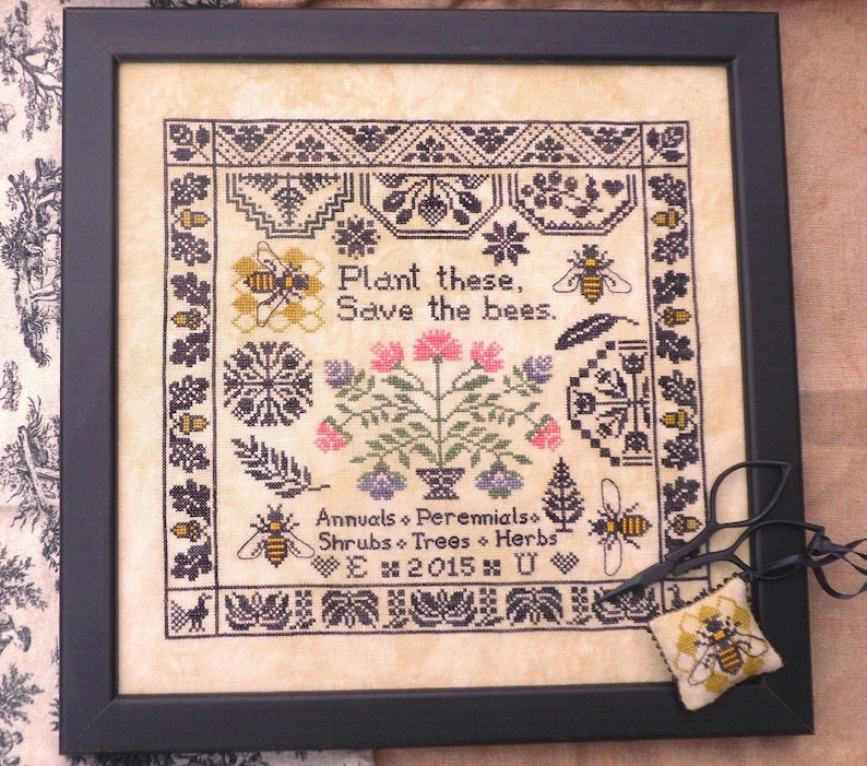 Puede incluir: Arte de punto de cruz enmarcado con un marco negro. El dise&ntilde;o presenta abejas, flores y el texto "Plant these, Save the bees". El texto adicional incluye "Annuals, Perennials, Shrubs, Trees, Herbs". Se incluye un peque&ntilde;o alfiletero con tem&aacute;tica de abejas y tijeras.