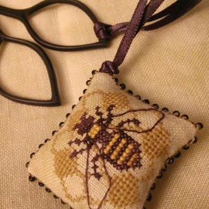 Puede incluir: Adorno de abeja hecho a mano con punto de cruz. El adorno cuadrado presenta un dise&ntilde;o detallado de abeja en tonos marr&oacute;n y dorado, con un borde de cuentas negras. Cuelga de una cinta morada oscura. El fondo es una tela beige claro.