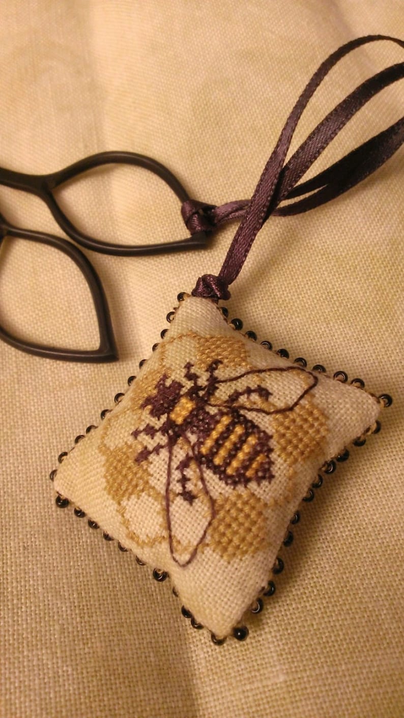 Puede incluir: Adorno de abeja hecho a mano con punto de cruz. El adorno cuadrado presenta un dise&ntilde;o detallado de abeja en tonos marr&oacute;n y dorado, con un borde de cuentas negras. Cuelga de una cinta morada oscura. El fondo es una tela beige claro.