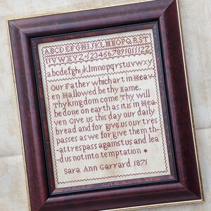 May include: A framed cross-stitch sampler with red embroidery thread on a cream-colored fabric. The sampler features the Lord's Prayer and the alphabet in a vintage font. The text reads "Our Father which art in heaven, hallowed be thy name...Thy kingdom come, Thy will be done on earth as it is in heaven. Give us this day our daily bread and forgive us our trespasses as we forgive them that trespass against us and lead us not into temptation. Sara Ann Garrard 1871."