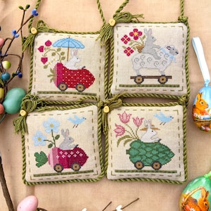 May include: Four embroidered cross-stitch ornaments featuring bunnies riding in various vehicles. The ornaments are framed with a green rope trim and have a tassel on each corner. The ornaments depict a bunny in a red car with a strawberry, a bunny in a green turtle shell, a bunny in a blue car with a strawberry, and two bunnies in a white wagon.