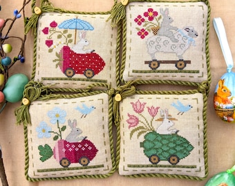 May include: Four embroidered cross-stitch ornaments featuring bunnies riding in various vehicles. The ornaments are framed with a green rope trim and have a tassel on each corner. The ornaments depict a bunny in a red car with a strawberry, a bunny in a green turtle shell, a bunny in a blue car with a strawberry, and two bunnies in a white wagon.