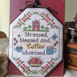 May include: A cross-stitch design with a red house, green trees, and a butterfly. The text reads "Stressed, blessed and coffee obsessed".