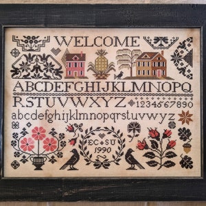 May include: Framed cross-stitch sampler with the word "WELCOME" at the top. The design includes houses, a pineapple, birds, flowers, and the alphabet. The frame is black and the sampler is on a beige background.