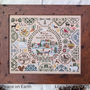 Op de afbeelding: Een kruissteek-sampler met een winters dorpstaferel en de tekst "Peace on Earth Goodwill to men." De sampler is ingelijst in een donkerbruin houten lijst.