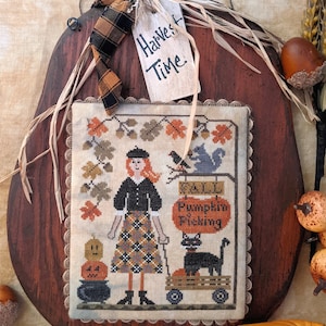 Peut inclure: Une broderie au point de croix sur une plaque en bois en forme de citrouille. La broderie représente une femme en robe noire et jupe à carreaux, un chat noir et une citrouille avec le texte "Fall Pumpkin Picking". La plaque est décorée d'un ruban à carreaux brun et noir et d'une étiquette qui dit "Harvest Time".