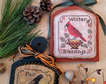 May include: Two wooden signs with cross-stitch designs. The top sign is red and features a cardinal with the words "Winter Blessings". The bottom sign is black and features a crow with the words "Fall Blessings".