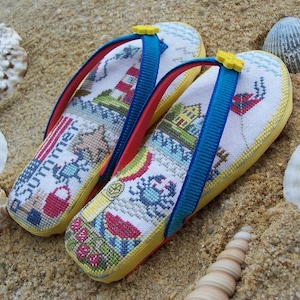 May include: A pair of yellow flip-flops with blue straps and a white canvas top featuring a summer-themed cross-stitch design. The design includes a lighthouse, a crab, a beach umbrella, a seashell, a starfish, and the word "Summer".