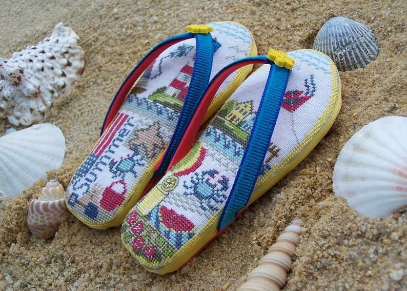 May include: A pair of yellow flip-flops with blue straps and a white canvas top featuring a summer-themed cross-stitch design. The design includes a lighthouse, a crab, a beach umbrella, a seashell, a starfish, and the word "Summer".
