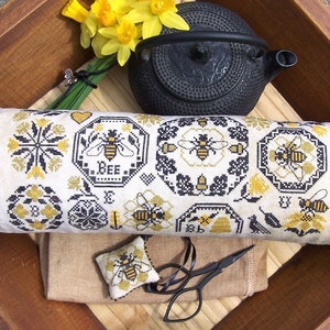 May include: A black and white embroidered cushion featuring a bee theme. The cushion is decorated with bees, honeycombs, and flowers. The cushion is resting on a wooden tray with a black teapot and a bunch of yellow daffodils.