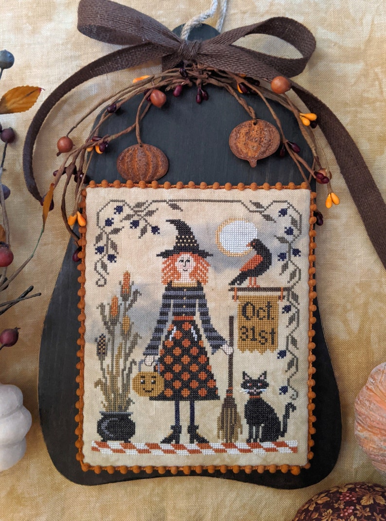 Op de afbeelding: Een kruissteekontwerp met een heks die een pompoenmandje en een bezem vasthoudt, met een zwarte kat en een vogel op een bord met de tekst "Oct 31st". Het ontwerp is ingelijst in een donkerhouten lijst met een bruin lint en herfstdecoraties.