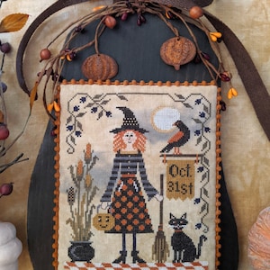 Pode incluir: Uma decoração de parede em ponto cruz com tema de Halloween. O design apresenta uma bruxa, um gato preto e um letreiro que diz "Oct 31st". A peça é emoldurada com um suporte de madeira escura e adornada com elementos decorativos.
