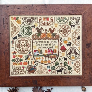 May include: A framed cross-stitch sampler with an autumnal theme. The design features an acorn with a house, a turkey, and a variety of other autumnal motifs. The text reads "Autumn is as joyful and sweet as an untimely end."