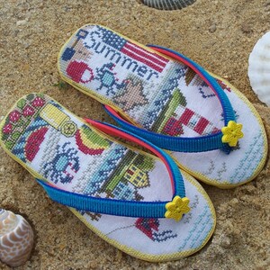 May include: A pair of yellow flip-flops with blue and red straps. The flip-flops are decorated with a cross-stitch design featuring a lighthouse, a crab, and the word "Summer".
