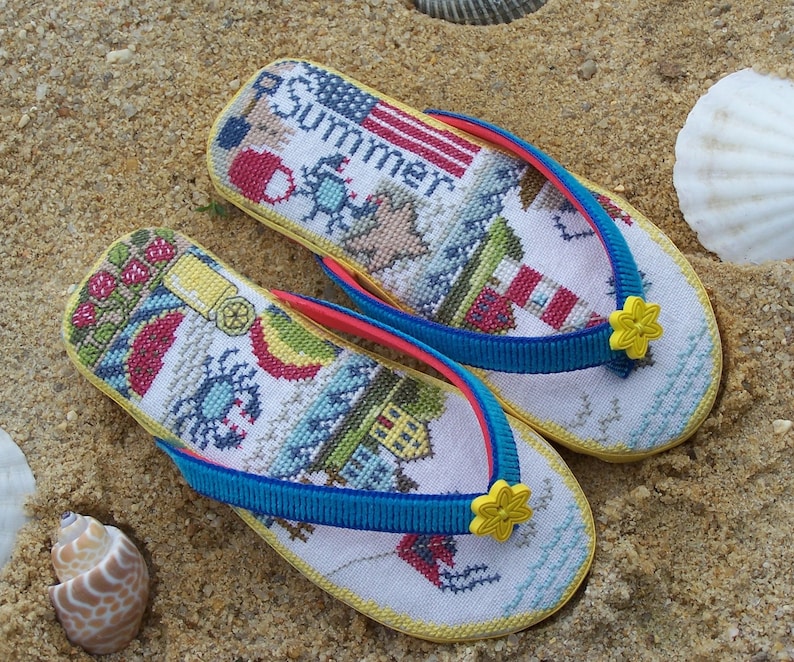 May include: A pair of yellow flip-flops with blue and red straps. The flip-flops are decorated with a cross-stitch design featuring a lighthouse, a crab, and the word "Summer".