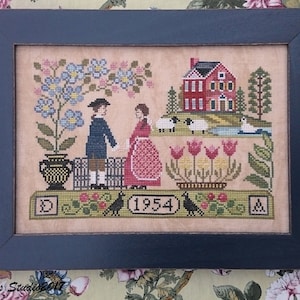Può includere: Un ricamo a punto croce incorniciato in una cornice blu scuro. Il disegno presenta una coppia in piedi davanti a una casa rossa con una staccionata bianca. Nella scena ci sono fiori, uccelli e pecore. L'anno 1954 è ricamato al centro in basso.