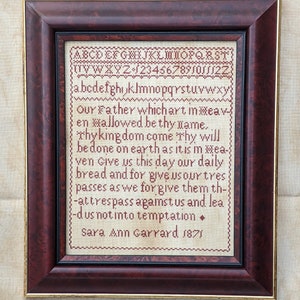 May include: Framed cross-stitch sampler with a dark brown wooden frame. The sampler features the alphabet, numbers, and a religious verse in red thread on a beige background. The text includes the name "Sara Ann Garrard 1871".