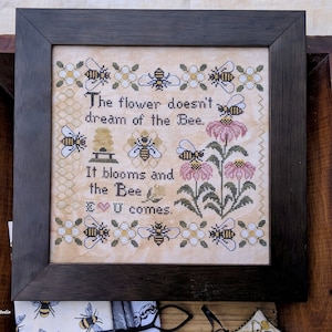 Op de afbeelding: Een borduurwerk met kruissteek, ingelijst in een donkerhouten lijst. Het borduurwerk toont een bijenkorf, bijen, bloemen en de tekst "The flower doesn't dream of the Bee. It blooms and the Bee U comes."