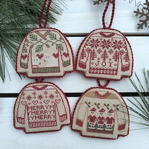 Può includere: Quattro addobbi natalizi fatti a mano a forma di maglioni con diversi design. I maglioni sono beige con bordi rossi e presentano vari motivi a tema natalizio, tra cui fiocchi di neve, alberi e le parole "Merry" e "Joy".