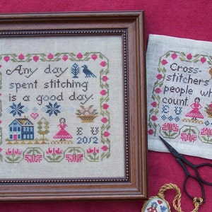 May include: Two framed cross-stitch embroidery pieces with a brown wooden frame. The first piece says "Any day spent stitching is a good day." The second piece says "Cross-stitchers are people who count." Both pieces feature floral designs and a small house.