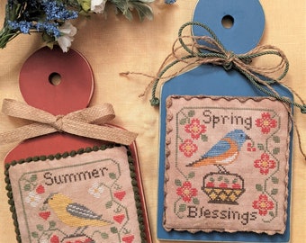 May include: Two wooden signs with embroidered designs. The top sign is blue with a bluebird and the text "Spring Blessings". The bottom sign is red with a yellow bird and the text "Summer Blessings".