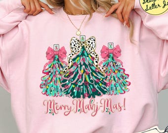 May include: A light pink sweatshirt featuring a graphic of three stylized Christmas trees decorated with mahjong tiles and large pink bows. The central tree has a leopard print bow. The text Merry Mahj-Mas! is printed in red cursive script.