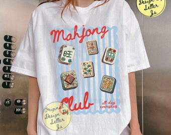 May include: A white t-shirt featuring a graphic print of six mahjong tiles arranged over a light blue vertical striped background. The red text reads Mahjong Club all day everyday. The shirt has short sleeves and a crew neckline design.