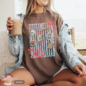May include: Brown t-shirt with a graphic featuring Mahjong tiles and the text "Cracks, Dots, Bams, Mahy Club". The design includes a red bow and blue striped background. The shirt is a comfortable, casual style.
