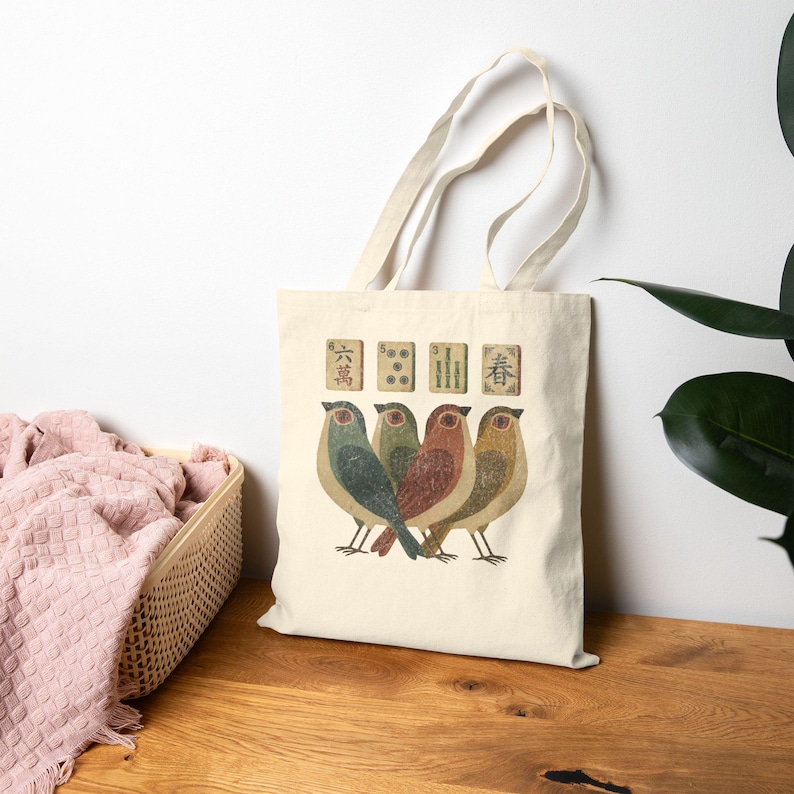 May include: A cream-colored canvas tote bag featuring a graphic print of four small birds standing in a row. Above the birds are four rectangular mahjong tiles, each displaying unique symbols and Chinese characters in muted, earthy tones.