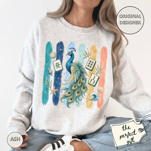 May include: Pale grey sweatshirt with a peacock illustration and vertical stripes in various colours. The peacock is depicted in blue and yellow, with a detailed tail. The design includes seashells, stars, and Chinese characters. The words "ORIGINAL DESIGNER" are in a circle.