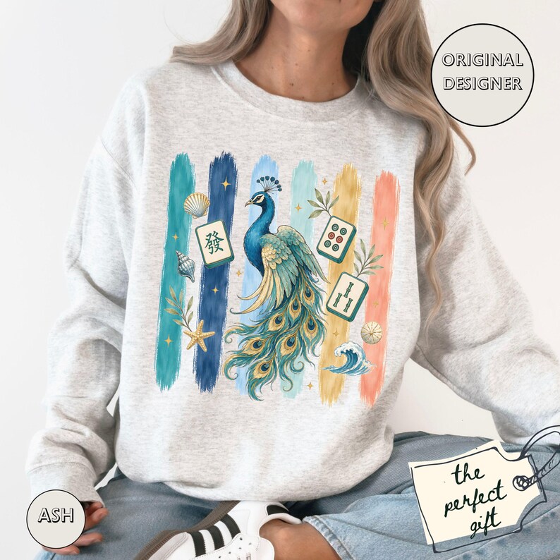 May include: Pale grey sweatshirt with a peacock illustration and vertical stripes in various colours. The peacock is depicted in blue and yellow, with a detailed tail. The design includes seashells, stars, and Chinese characters. The words "ORIGINAL DESIGNER" are in a circle.