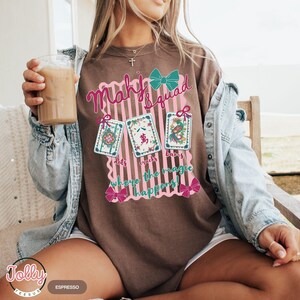 May include: Brown t-shirt with a graphic that says "Mahj Squad" and "where the magic happens." The graphic includes three playing cards and pink bows. The shirt is a comfortable, casual style.