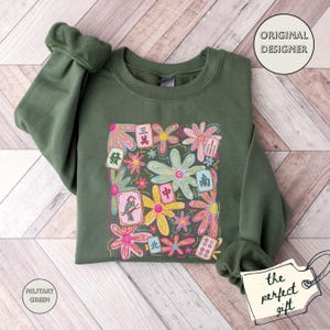 May include: A military green crewneck sweatshirt featuring a central graphic print of colorful hand-drawn style flowers interspersed with various mahjong tiles, including tiles with Chinese characters and a bird illustration, on the front chest.