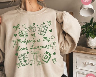 May include: Sand-colored sweatshirt with a green graphic that reads "Mahjong is My Love Language." The design includes Mahjong tiles, hearts, bows, and the word "Joker." The sweatshirt is a casual, comfortable style.