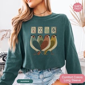 May include: A blue spruce long-sleeve crewneck sweatshirt featuring a graphic print of four birds standing in a row. Above the birds are four mahjong tiles displaying the characters for six, five, three, and spring in a vintage-inspired style.