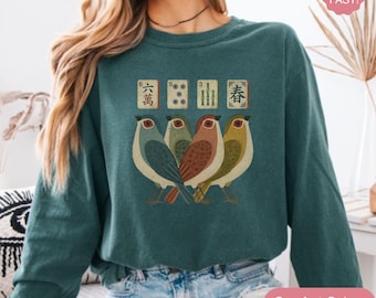 May include: A blue spruce long-sleeve crewneck sweatshirt featuring a graphic print of four birds standing in a row. Above the birds are four mahjong tiles displaying the characters for six, five, three, and spring in a vintage-inspired style.