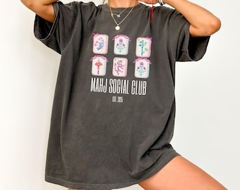 May include: Dark grey t-shirt featuring a graphic of six floral Mahjong tiles. The text "MAHJ SOCIAL CLUB" is printed below the tiles, with "EST. 2025" underneath. The shirt has short sleeves and a relaxed fit.