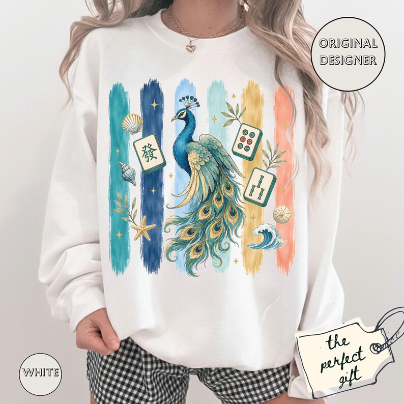 May include: White sweatshirt with a peacock design and vertical stripes in various colours. The peacock has blue and gold feathers, and the design includes Mahjong tiles, seashells, and a wave. The words "Original Designer" are in a circle.