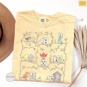 May include: Butter-colored t-shirt with a whimsical illustration of birds and Mahjong tiles on branches. The design includes a Comfort Colors label and the words "Original Designer". A delicate necklace with a star pendant is also visible.